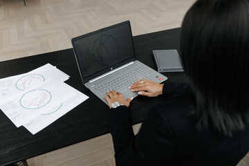 An astrologer girl sits at a black wooden desk and holds a natal chart with cards with the signs of the zodiac. The computer and day planner are in the background. 