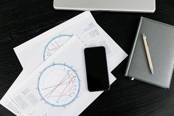 The notepad for writing down ideas and the astrological natal birth chart lie on a black background. A computer keyboard and mouse next to it and a phone. Astrological consultation idea.