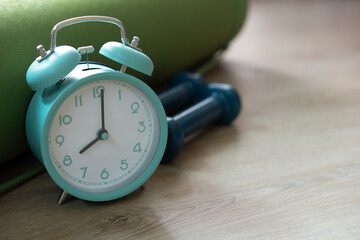 Time for exercising clock and dumbbell with green yoga mat on wooden table background, sport and healthy concept