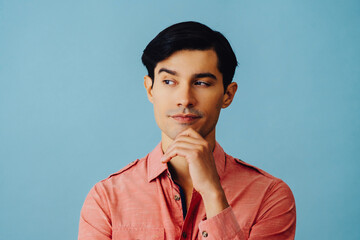 Headshot thoughtful handsome young adult latino man with hand on chin black hair and pink shirt over blue background looking away at copy space studio shot - obrazy, fototapety, plakaty