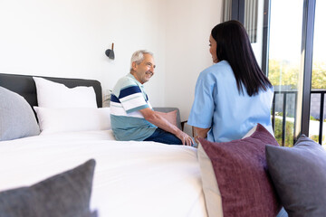 Biracial female health worker and caucasian senior man sitting on the bed at home