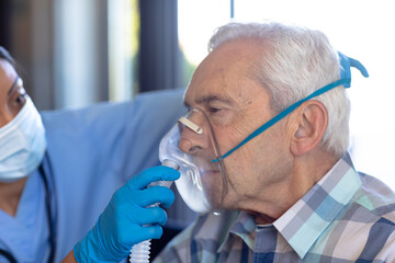 Biracial female health worker helping caucasian senior man to use oxygen mask at home