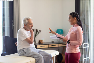 Biracial female physiotherapist with clipboard discussing with caucasian senior man at home