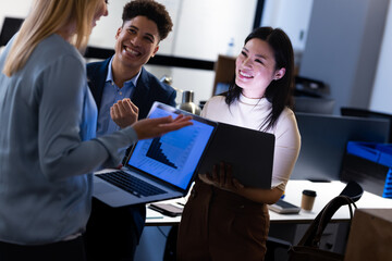 Happy multiracial colleagues analyzing charts over laptops while working late in office at night