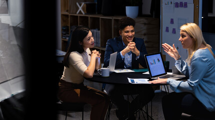 Multiracial female financial advisor with laptop explaining data to coworkers in meeting at night