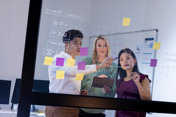 Multiracial coworkers brainstorming over data and adhesive notes on glass wall in office at night