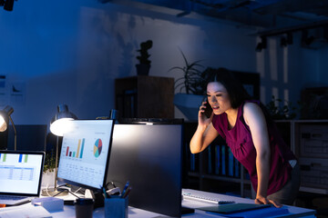 Asian female professional talking over mobile phone while looking at computer in office at night