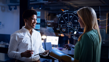 Smiling multiracial coworkers with report and digital pc standing by glass wall in office at night