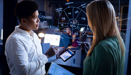 Multiracial colleagues with reports analyzing and brainstorming by glass wall in office at night