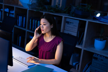 High angle view of asian businesswoman talking on smartphone and using computer at desk in office