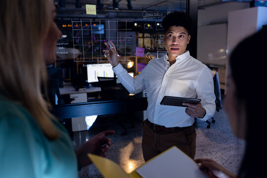 Multiracial businessman with digital pc explaining data on glass wall to female coworkers in office