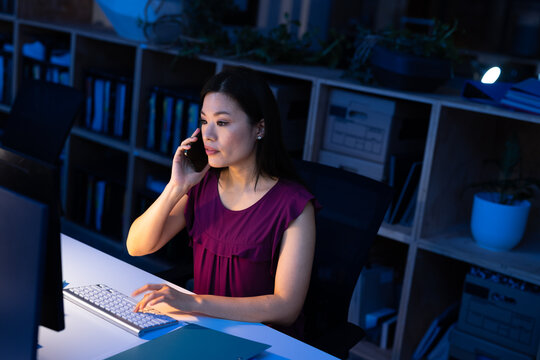 High angle view of asian businesswoman talking on smartphone and using computer at desk in office