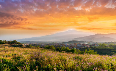 view from hill with golden grass and green bushes to a valley town with majectic mountains and scenic cloudy sunset on background