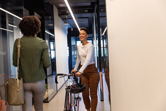 Smiling biracial businesswoman with bicycle by african american colleague in corridor of office