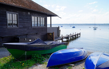 a scenic view of lake Ammersee in Germany with a boat house and a couple of sailing boats resting on the water on a sunny summer day with light white clouds (Herrsching, Bavaria, Germany)	