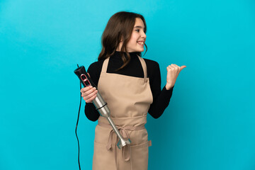 Little girl using hand blender isolated on blue background pointing to the side to present a product