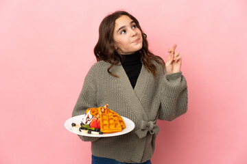 Little girl holding waffles isolated on pink background with fingers crossing and wishing the best