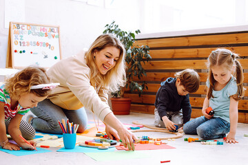 Smiling woman educator playing with children sitting on floor in kindergarten. Kids folding blocks and drawing with colored pencils in playroom closeup. Toys for kindergartners developing intelligence - obrazy, fototapety, plakaty
