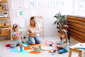 Woman educator play with children sitting on floor in kindergarten. Kids folding blocks and drawing...