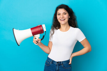 Teenager Russian girl isolated on blue background holding a megaphone and smiling
