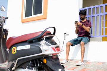 Young man using mobile phone near charging point during recharging of ev or electric scooter -...