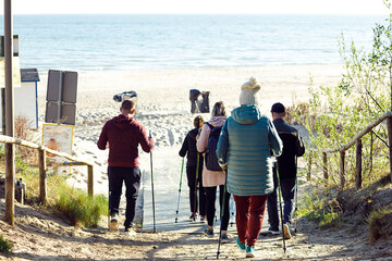 Rearview of people group campers with backpacks making Nordic walking with professional sticks going down to sea beach