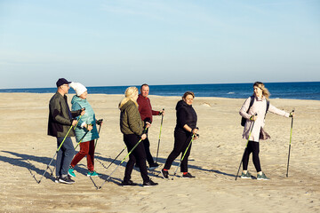 Leader sportswoman, trainer and people group joying Scandinavian walking with sticks at sandy beach near sea. Copy space - obrazy, fototapety, plakaty
