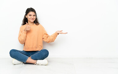 Teenager Russian girl sitting on the floor holding copyspace imaginary on the palm to insert an ad and with thumbs up