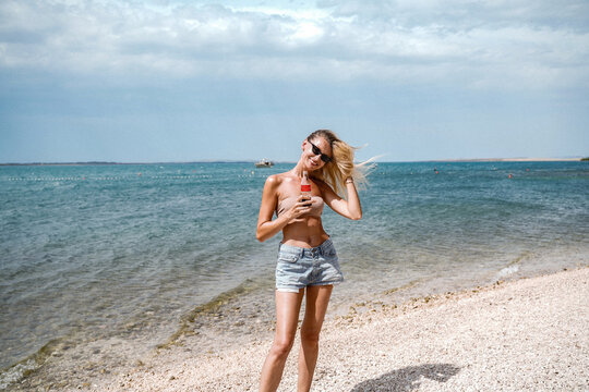Summer photo of blonde happy woman relaxing on the beach, smiling. Girl with fit tan body wearing short jeans.