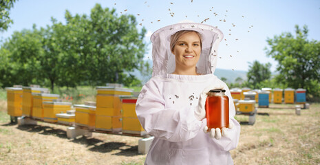 Young female bee keeper in a uniform holding a jar of honey on apiary