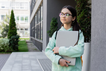 Preteen schoolchild in eyeglasses holding laptop on urban street.