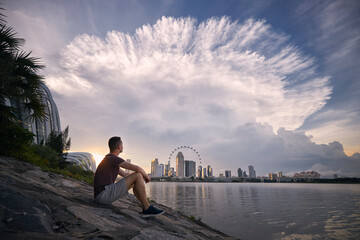 Young man sitting on waterfront and looking at dramatic clouds of thunderstorm above urban skyline,...
