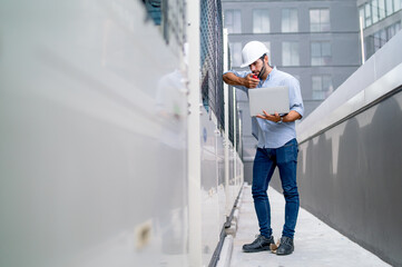 Wide shot of Caucasian engineer or technician worker use laptop to work in way on terrace of construction site with high building as background.