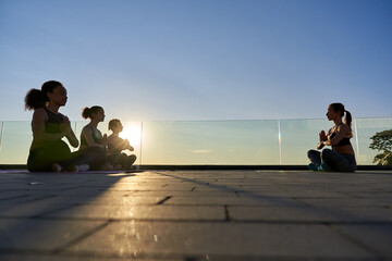 Diverse ethnicity female silhouettes and yoga instructor sitting on ground outdoor in yoga poses...