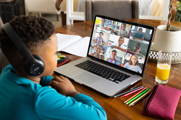 African american boy student wearing headphones and attending online class over laptop at home