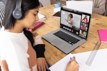 Biracial teenage girl wearing headphones writing while following professor teaching in online class - Powered by Adobe