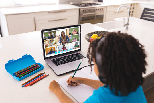 Biracial boy with colored pencils on table writing in book while studying online over laptop at home - Powered by Adobe