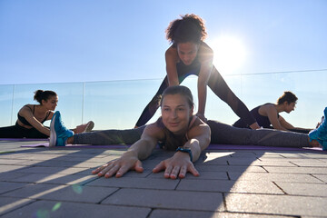 Fit sporty afro american trainer coach helping flexible caucasian woman friend during group yoga class outdoor. Fit female yoga teacher teaching lady bending exercise on pilates fitness class outside.