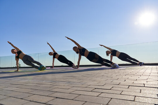 Fit healthy sporty young adult multiethnic women friends silhouettes stand in yoga pose side plank at active outdoor group class do fitness exercise stretching in balance together at retreat workout.