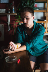 Portrait of young man looking worried smoking cigarette at pub or bar. Nicotine addicted person with bad habits, sitting lonely and thoughtful