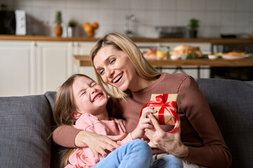 Happy mom receiving Mothers Day gift box hugging daughter at home. Cute kid girl laughing, having fun making present greeting mum on spring holiday while mommy hugging child sitting on sofa.