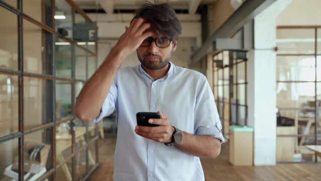 Indian business man using cellphone apps working in office. Professional ethnic businessman holding mobile phone checking finance banking application technology while working in hall.