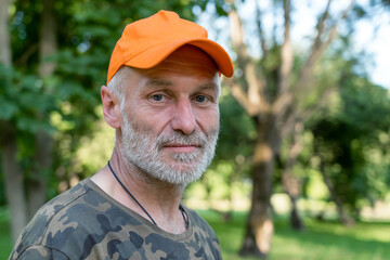 Close up portrait of sad mature man wearing orange cap looking at camera, standing in park...