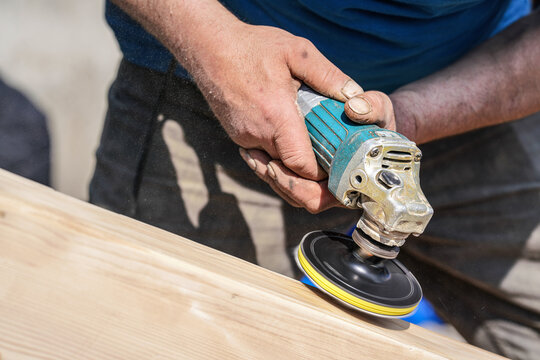 Man polishing wooden chest with old angle grinder during sunny day, closeup detail to hands without gloves, fine wood dust flying in air