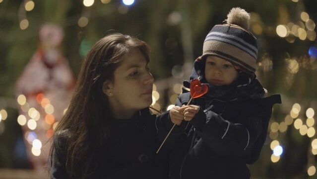 Little boy kiss mother lips close Christmas tree, heart shape lollipop