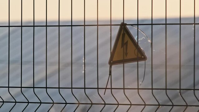 High voltage sign on a wire fence of the solar plant. Yellow triangular sign lightning danger high voltage. Warning electricity safety symbol of the power plant. Sign of the danger of electric shock.