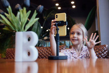 Cute little caucasian girl with blonde hair in trendy casual clothes using mobile phone in modern...