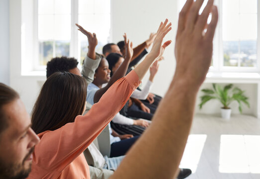 Happy diverse multiracial male female audience raising hands to ask speaker questions. Young mixed race multiethnic men and women sitting in row with hands up after interesting lecture or masterclass