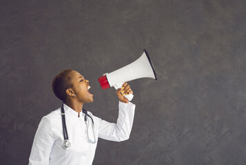 Angry young African American woman doctor on black studio background shout yell in loudspeaker. Mad furious ethnic female GP make announcement in speaker. Medicine, attention, healthcare concept. - Powered by Adobe