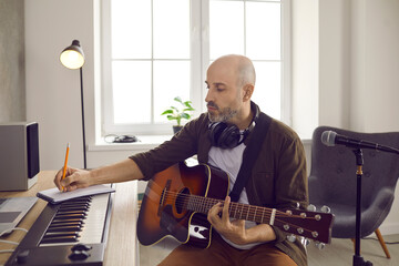 Focused male musician writes in notebook while sitting with guitar in music studio. Caucasian man working with synthesizer, headphones and guitar picks up lyrics for future song and writes down notes. - Powered by Adobe